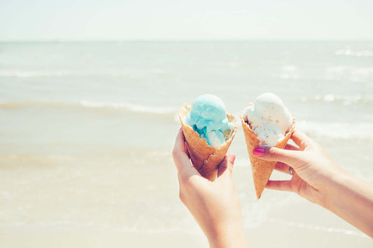 Two hands holding cones of sea salt ice cream by the beach, highlighting the DIY: Sea Salt Ice Cream Recipe.