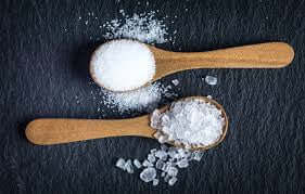 Two wooden spoons with different types of salt against a dark background, showcasing their texture and color.